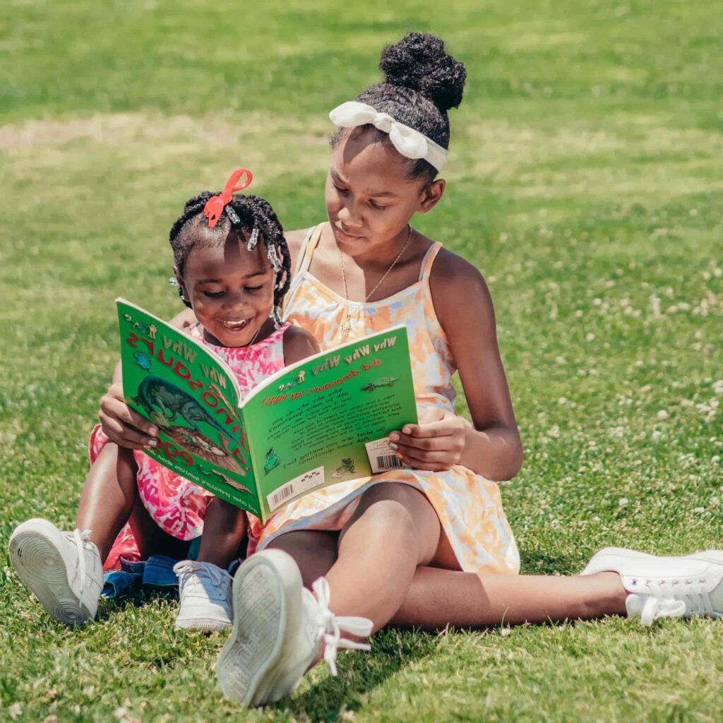 Two young girls sitting on grass reading a green book together