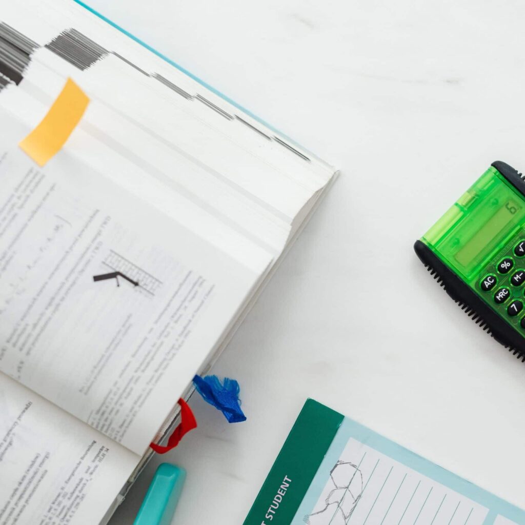 Open textbook with graph, green calculator, and teal student notepad on white surface