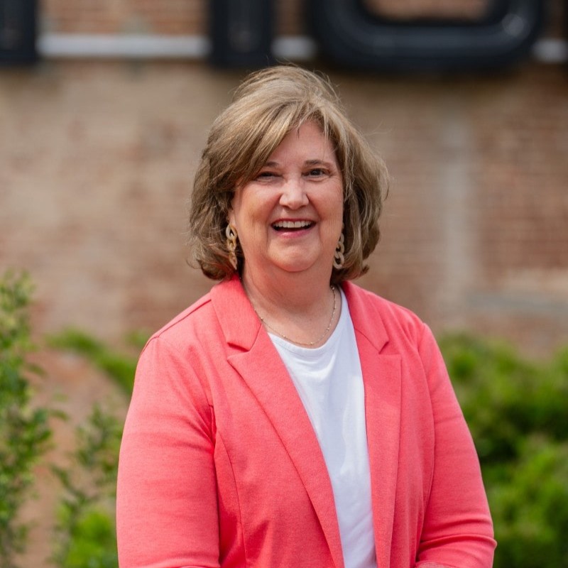 Woman smiling in coral blazer and white shirt, standing outdoors in front of brick wall.
