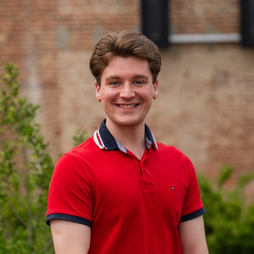 Smiling man in red polo shirt standing outdoors near a brick wall.