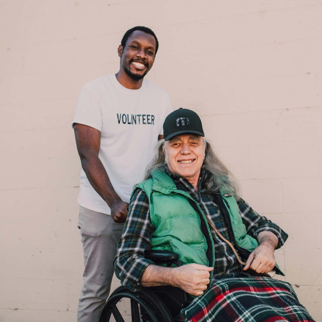 Volunteer pushing smiling man in wheelchair wearing green vest and plaid blanket.