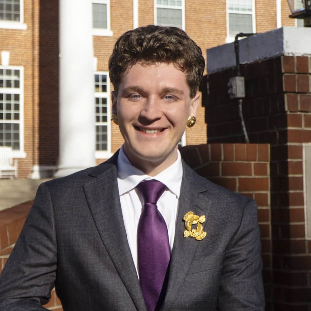 Eben Leigh, Community Impact Consultant for United Way of Danville-Pittsylvania County, stands outdoors in front of Averett College, smiling in a gray suit with a purple tie and gold accessories, leaning on a brick and white stone wall in bright sunlight.